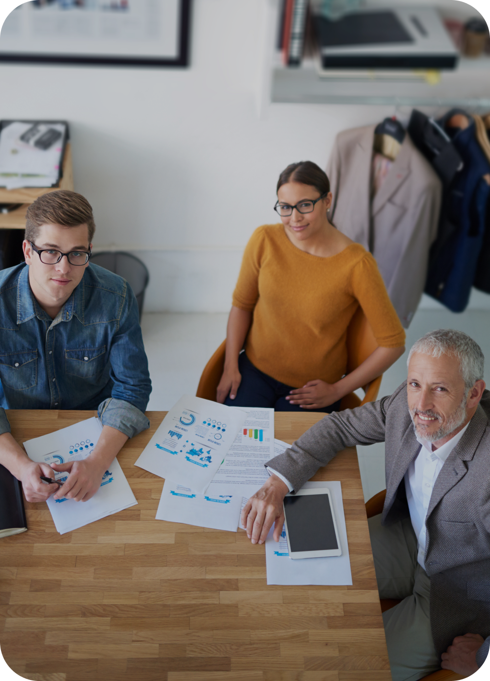 Tři lidé sedí u stolu v kanceláři, na stole jsou papíry s grafy, tablet a blok