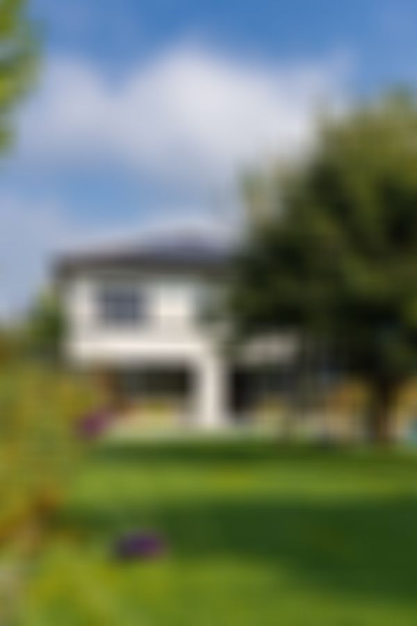 Modernes zweistöckiges Haus mit großen Glasfenstern, weiß-grauer Fassade, üppig grünem Garten und einem Baum unter teilweise bewölktem Himmel.