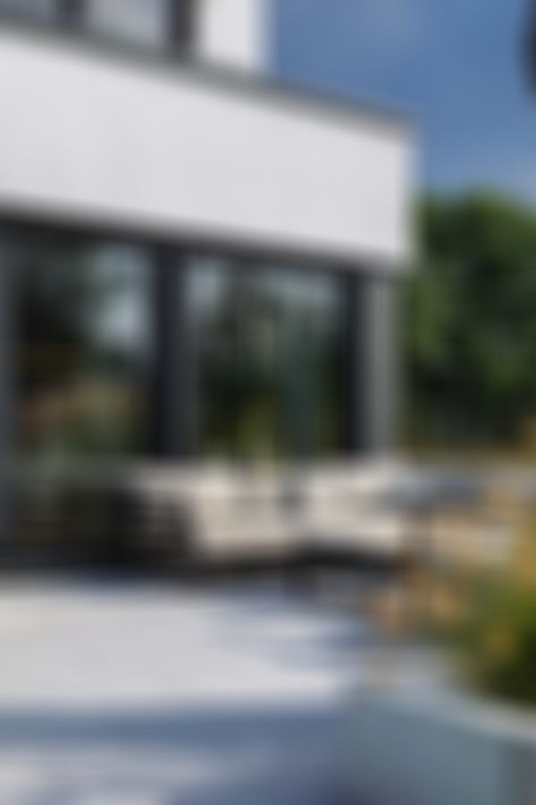 Moderne Terrasse mit beigem Sofa, dunklem Sonnenschirm und grünen Pflanzen. Große Glastüren führen in ein Esszimmer mit einer Hängeleuchte.