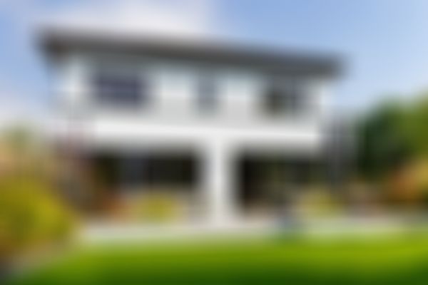 Modernes zweistöckiges Haus mit großen Fenstern, Sitzgelegenheiten auf der Terrasse und üppigem Garten. Ein Hund ruht sich auf der Terrasse aus, umgeben von üppigem Grün.