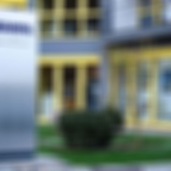 Modernes Gebäude mit gelb umrahmten Fenstern, daneben ein silbernes Schild mit der Aufschrift „KAMPA K3 Bauinnovationszentrum“, umgeben von Grünflächen.