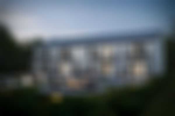 Modernes Mehrfamilienhaus in der Abenddämmerung mit beleuchteten Fenstern, Balkonen und einer runden Terrasse mit Feuerstelle und Stühlen, umgeben von Grünflächen.