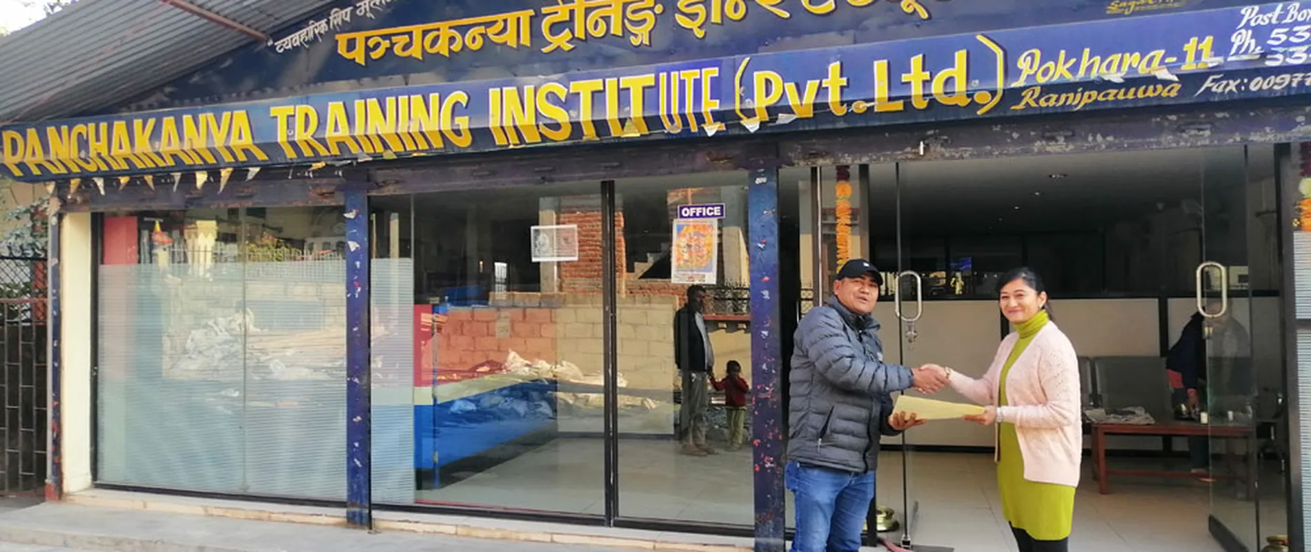 Two people shake hands in front of Panchakanya Training Institute. The building has large windows and a prominent sign above.