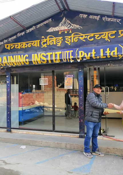 Two people shake hands in front of Panchakanya Training Institute. The building has large windows and a prominent sign above.