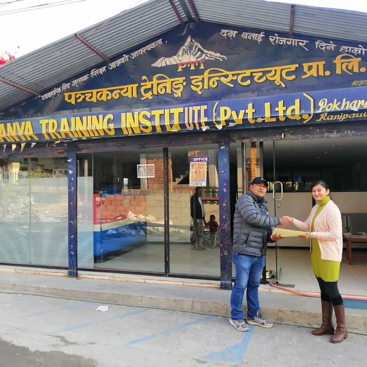 Two people shake hands in front of Panchakanya Training Institute. The building has large windows and a prominent sign above.