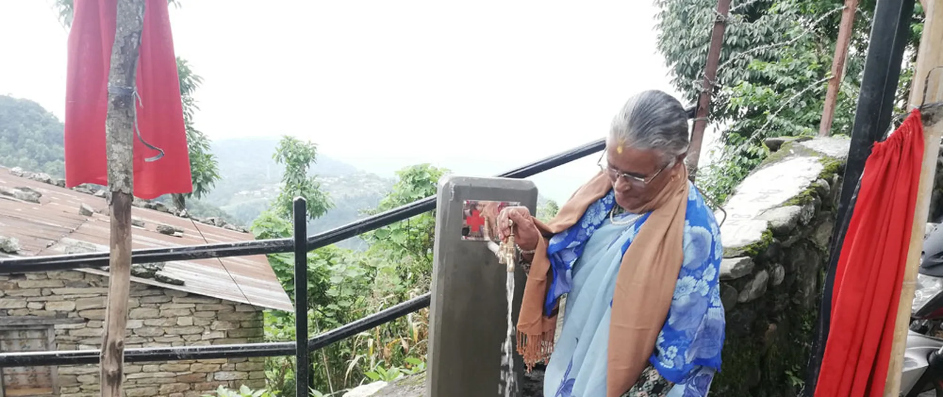 Elderly woman in a blue sari fills a copper pot with water from an outdoor tap, surrounded by greenery and stone buildings.