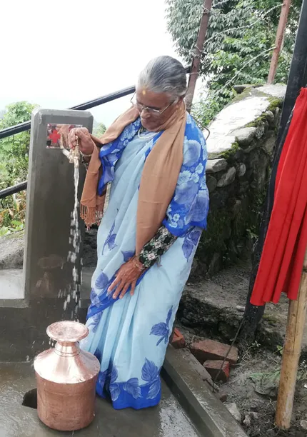 Elderly woman in a blue sari fills a copper pot with water from an outdoor tap, surrounded by greenery and stone buildings.