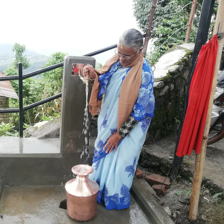 Elderly woman in a blue sari fills a copper pot with water from an outdoor tap, surrounded by greenery and stone buildings.