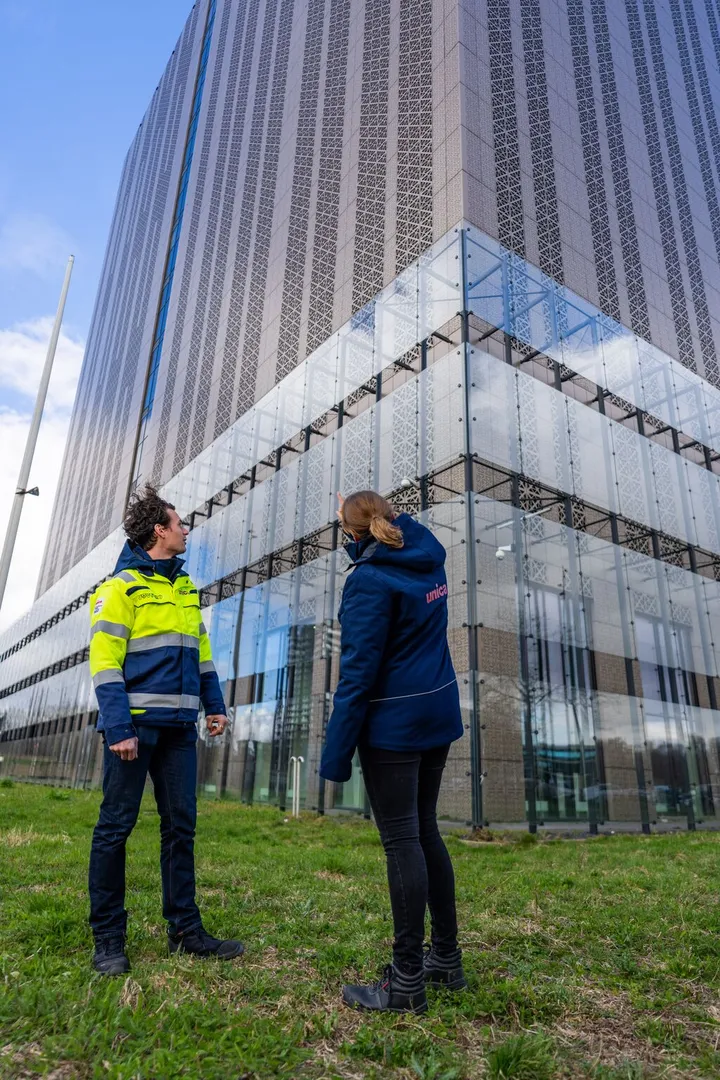 Two people in uniforms stand on grass, looking up at a modern glass and patterned facade skyscraper against a blue sky.