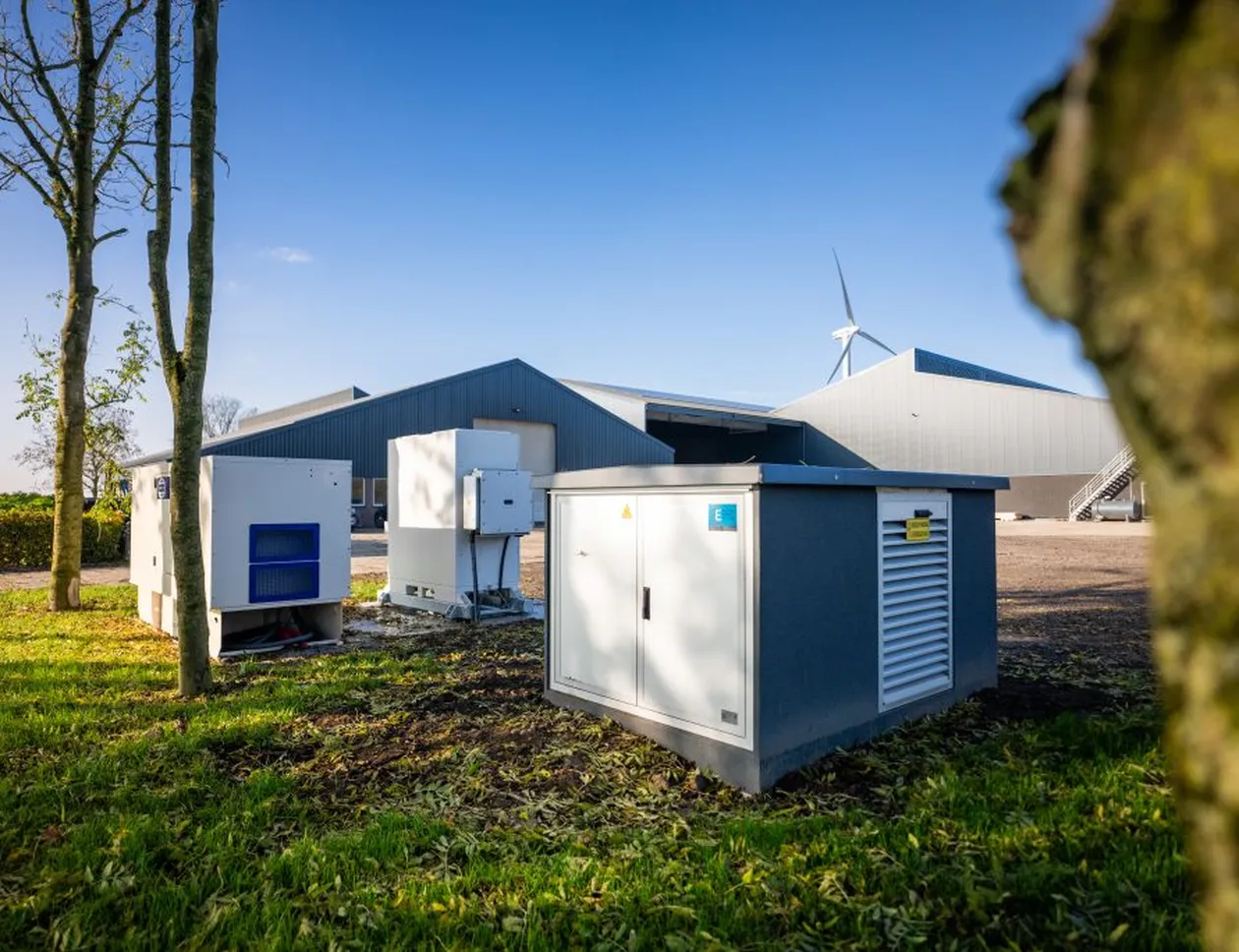 A farm with grey buildings, green lawn, and equipment. Wind turbines are visible in the background under a clear blue sky.