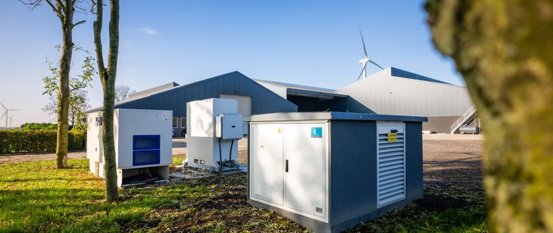 A farm with grey buildings, green lawn, and equipment. Wind turbines are visible in the background under a clear blue sky.