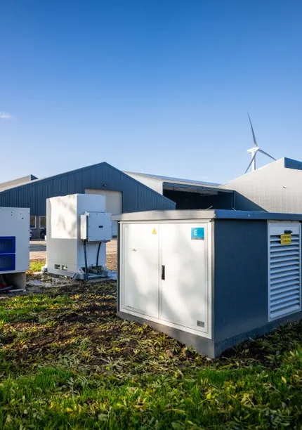 A farm with grey buildings, green lawn, and equipment. Wind turbines are visible in the background under a clear blue sky.