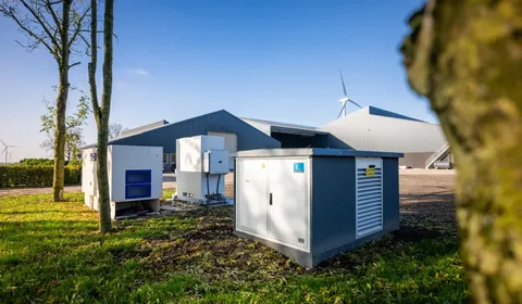 A farm with grey buildings, green lawn, and equipment. Wind turbines are visible in the background under a clear blue sky.