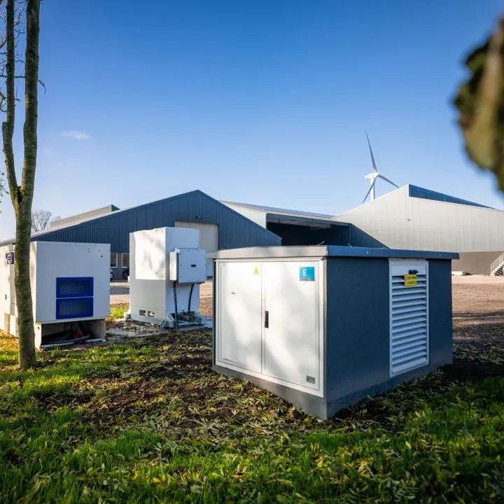 A farm with grey buildings, green lawn, and equipment. Wind turbines are visible in the background under a clear blue sky.