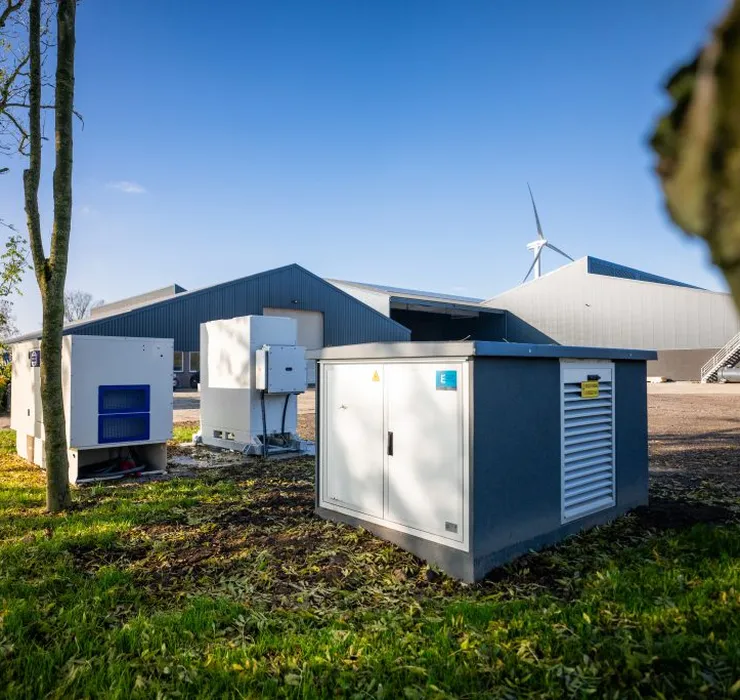A farm with grey buildings, green lawn, and equipment. Wind turbines are visible in the background under a clear blue sky.