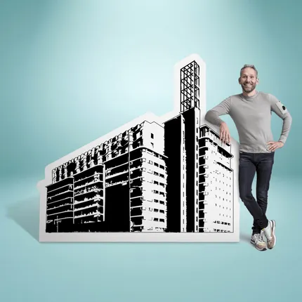 Man in gray sweater and sneakers leans on a cutout illustration of a modern building against a light blue background.