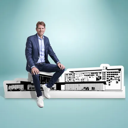 A man in a blue suit sits on a model of a modern building against a light blue background.
