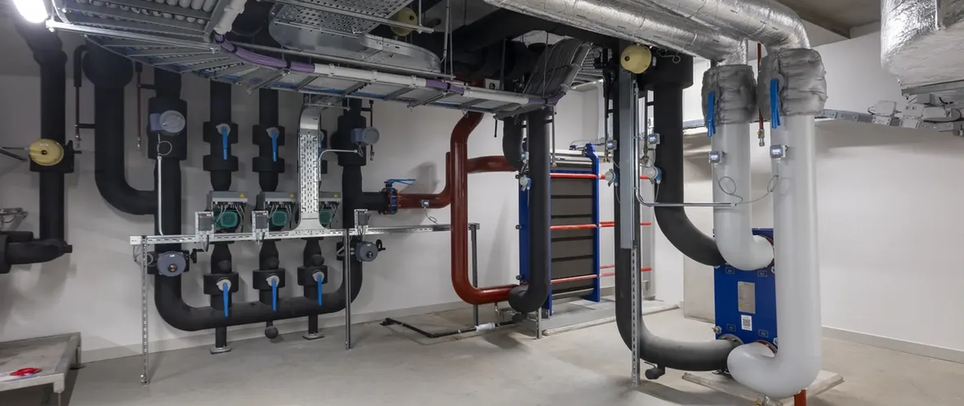 A clean, modern basement with exposed pipes, ducts, and industrial equipment, featuring insulated pipes and a neat arrangement on the walls.