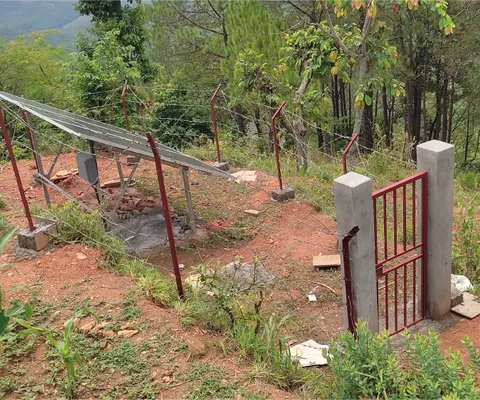A small solar panel installation on a hill, surrounded by a fence and vegetation, with distant forested mountains in the background.