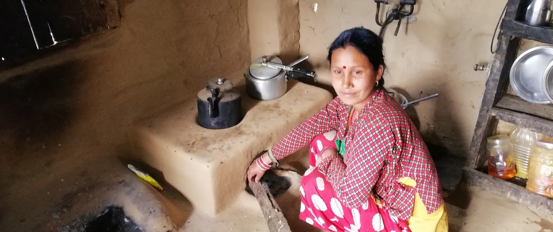 Woman in colorful attire kneels by a mud stove, adding wood, in a rustic kitchen with cooking pots and shelves.