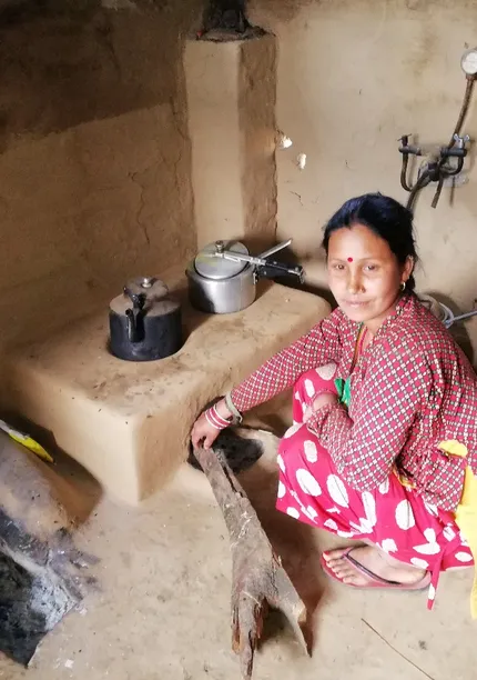 Woman in colorful attire kneels by a mud stove, adding wood, in a rustic kitchen with cooking pots and shelves.