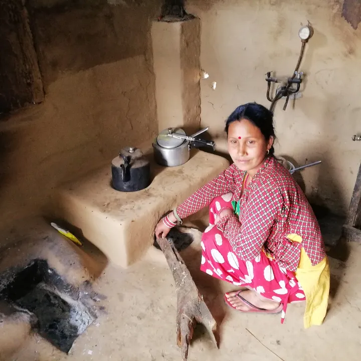 Woman in colorful attire kneels by a mud stove, adding wood, in a rustic kitchen with cooking pots and shelves.