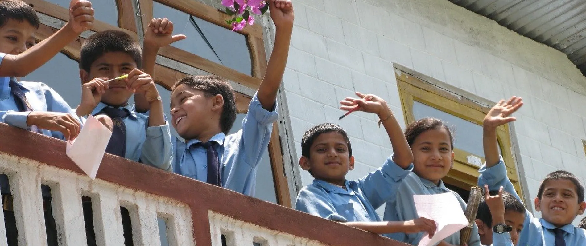 Children in school uniforms wave cheerfully from a balcony, holding flowers and papers, with a white building in the background.