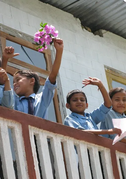 Children in school uniforms wave cheerfully from a balcony, holding flowers and papers, with a white building in the background.