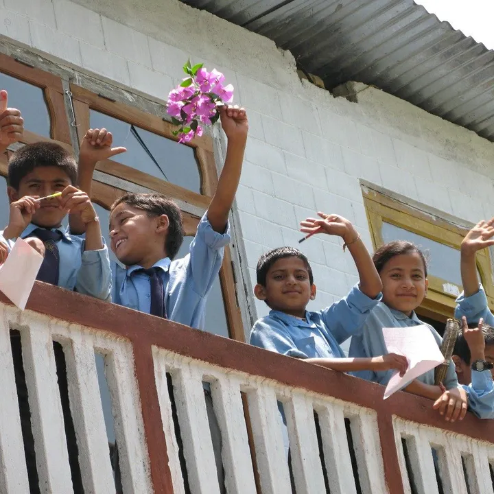 Children in school uniforms wave cheerfully from a balcony, holding flowers and papers, with a white building in the background.
