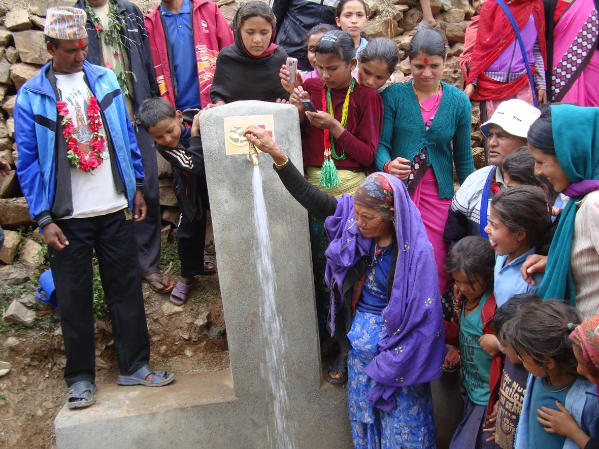 A group of people gather around a water tap outdoors. An elderly woman is filling a container with water. Some are dressed in traditional clothing.