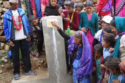 A group of people gather around a water tap outdoors. An elderly woman is filling a container with water. Some are dressed in traditional clothing.