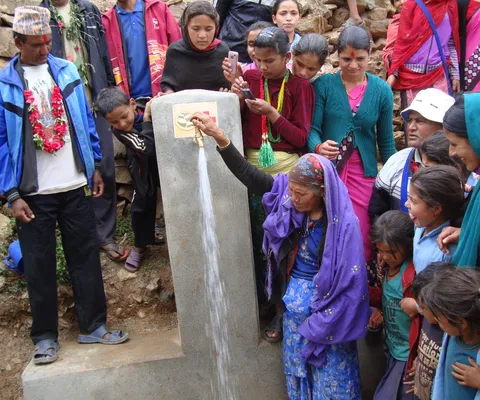 A group of people gather around a woman using a public water tap, built into a stone structure, in a rural setting.