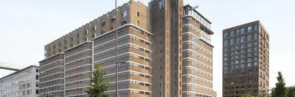 A modern, multi-story building complex with unique architectural features, large windows, and surrounding trees, set against a clear sky.