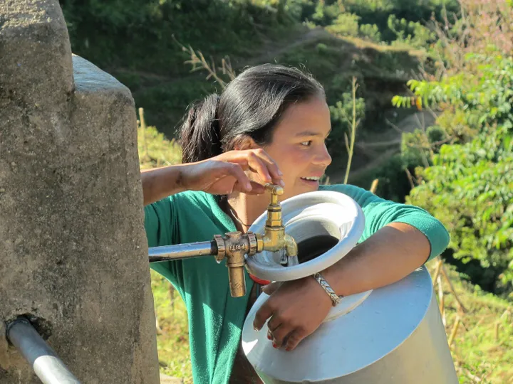 A woman fills a large metal container with water from a tap, set against a backdrop of greenery and sunny outdoor surroundings.