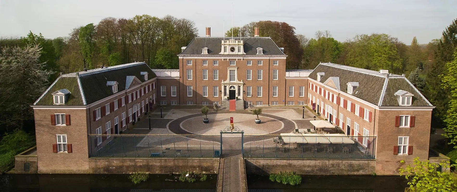 Aerial view of a historic brick mansion with two wings, a central courtyard, a circular driveway, and surrounded by trees and a water-filled moat.