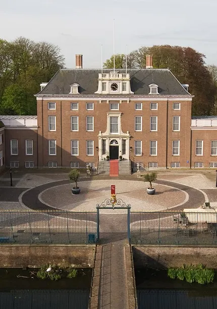 Aerial view of a historic brick mansion with two wings, a central courtyard, a circular driveway, and surrounded by trees and a water-filled moat.