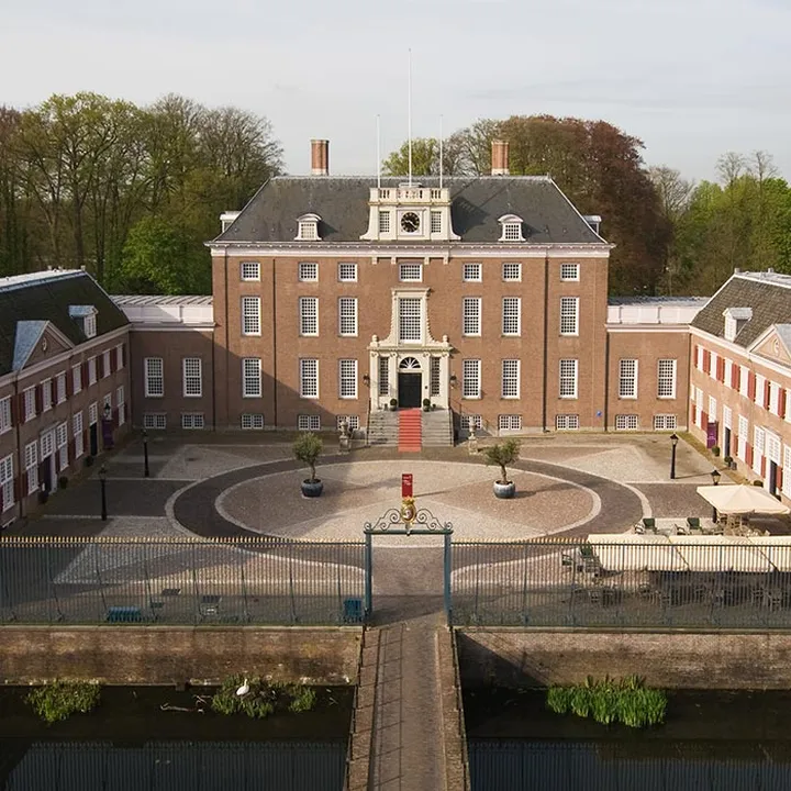 Aerial view of a historic brick mansion with two wings, a central courtyard, a circular driveway, and surrounded by trees and a water-filled moat.