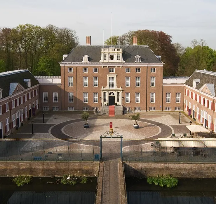 Aerial view of a historic brick mansion with two wings, a central courtyard, a circular driveway, and surrounded by trees and a water-filled moat.