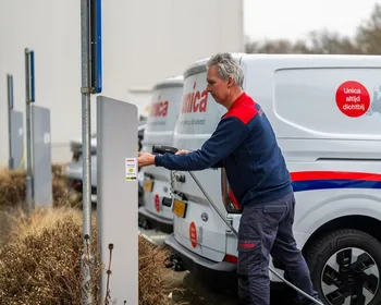 A man in a blue and red uniform plugs a charging cable into a white van with company branding in a parking area.