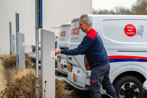 A man in a blue and red uniform plugs a charging cable into a white van with company branding in a parking area.