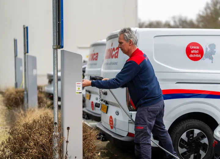 A man in a blue and red uniform plugs a charging cable into a white van with company branding in a parking area.
