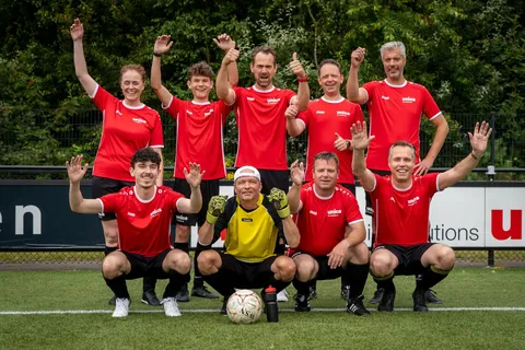 A soccer team in red jerseys and black shorts poses on a field, smiling with raised arms. A goalkeeper in yellow crouches in front.
