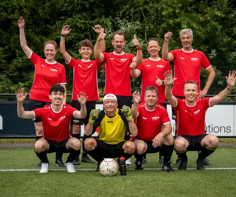 A soccer team in red jerseys and black shorts poses on a field, smiling with raised arms. A goalkeeper in yellow crouches in front.