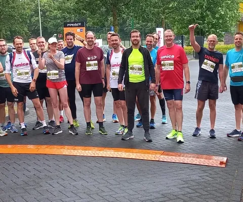 A group of runners poses at the starting line on a paved path, surrounded by trees and parked cars. Some raise their arms in excitement.