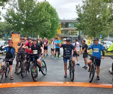 Cyclists and runners gather at a starting line in a park, wearing athletic gear and helmets, with trees and parked cars in the background.
