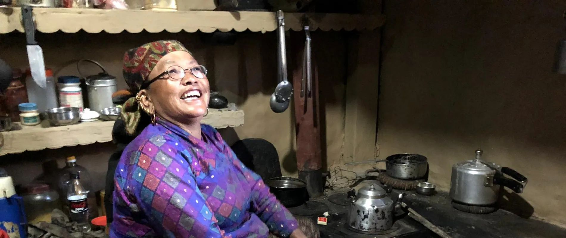 An elderly woman in traditional clothing sits by a stove, smiling, in a cozy kitchen with shelves of pots and utensils.