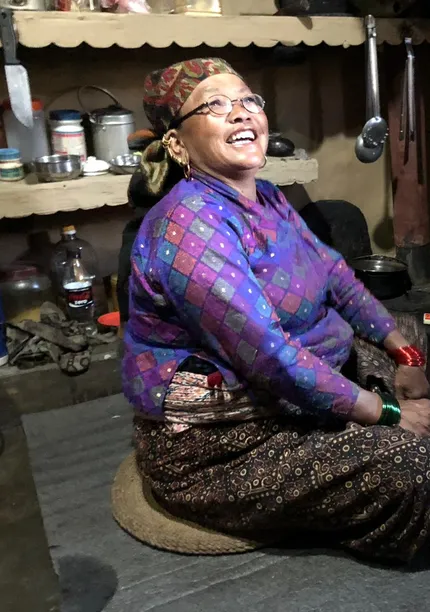 An elderly woman in traditional clothing sits by a stove, smiling, in a cozy kitchen with shelves of pots and utensils.