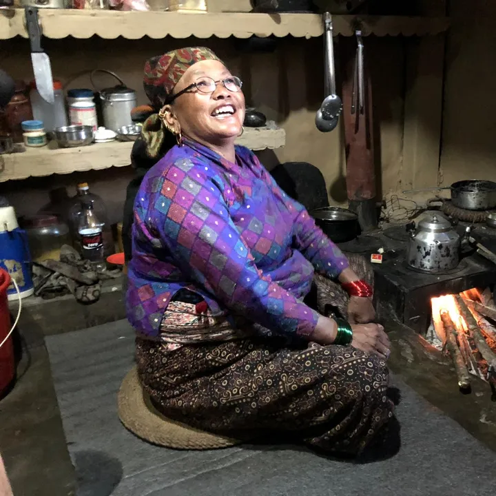 An elderly woman in traditional clothing sits by a stove, smiling, in a cozy kitchen with shelves of pots and utensils.