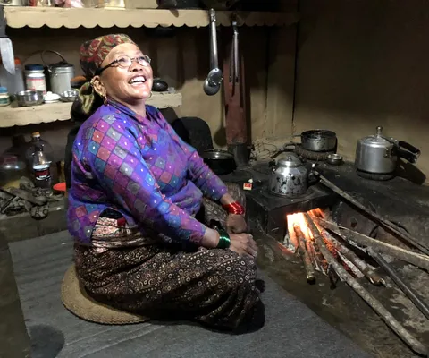 A woman in traditional attire smiles while sitting by a wood-burning stove in a rustic kitchen with shelves of utensils and cookware.