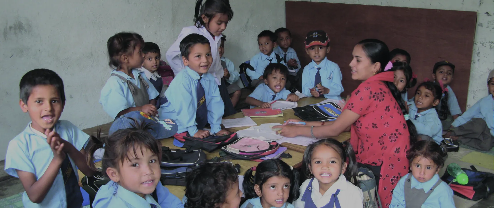 A group of young children in school uniforms gather around a table with their teacher in a classroom setting, smiling and engaged.